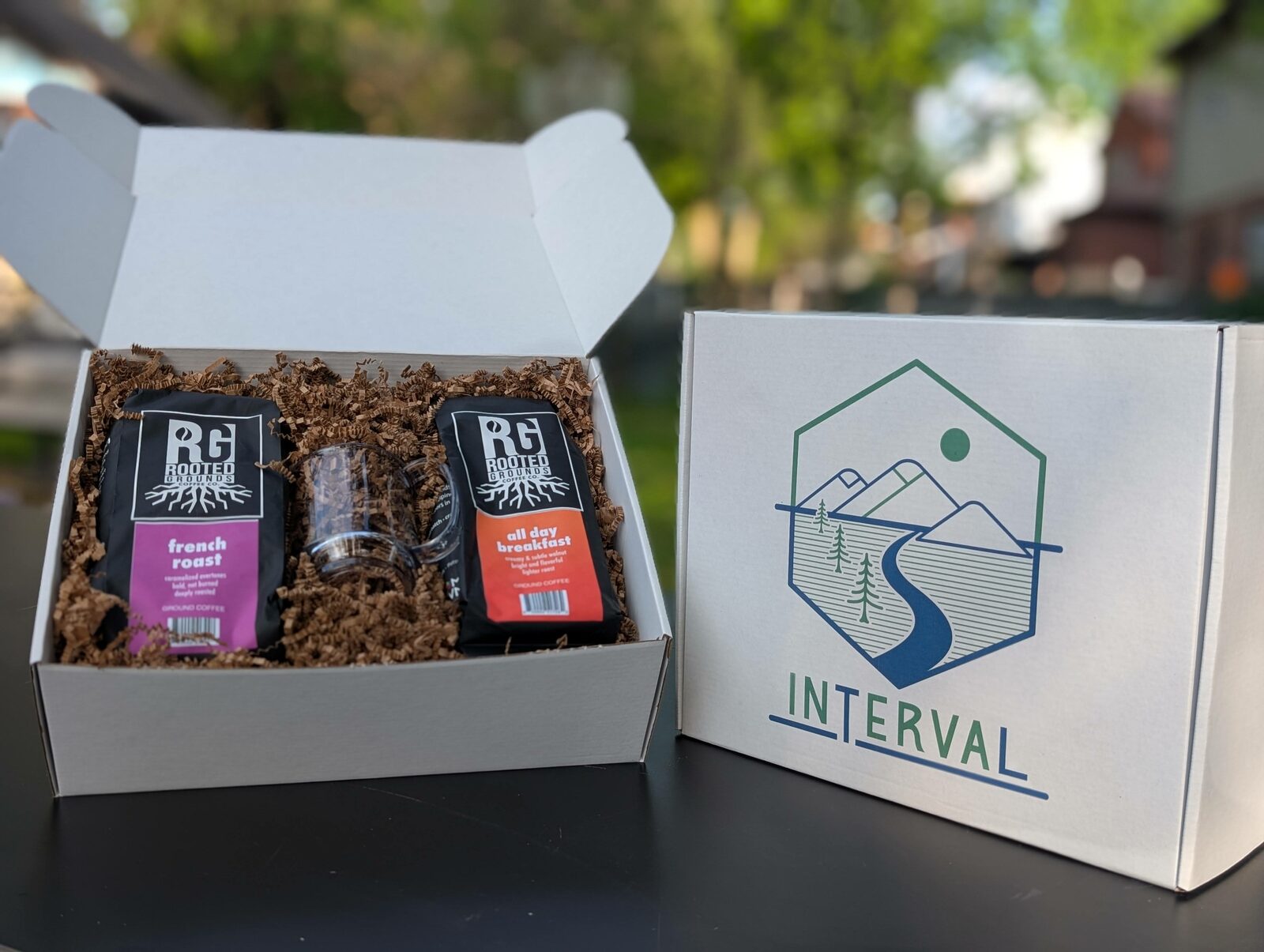 A full Interval Gift Box arranged on a wood table with French roast and breakfast blend coffee bags and a clear acrylic mug