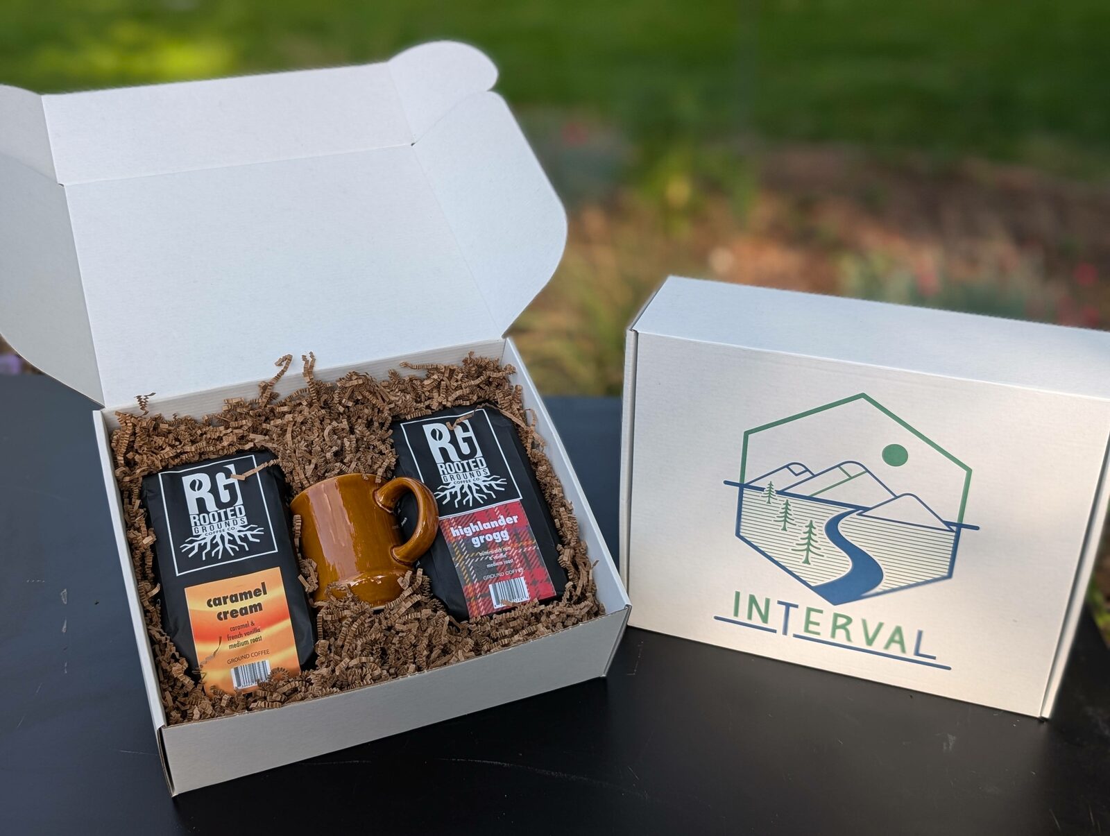 An open Interval gift box with two bags of Rooted Grounds coffee and a warm amber ceramic mug, set beside the closed branded box